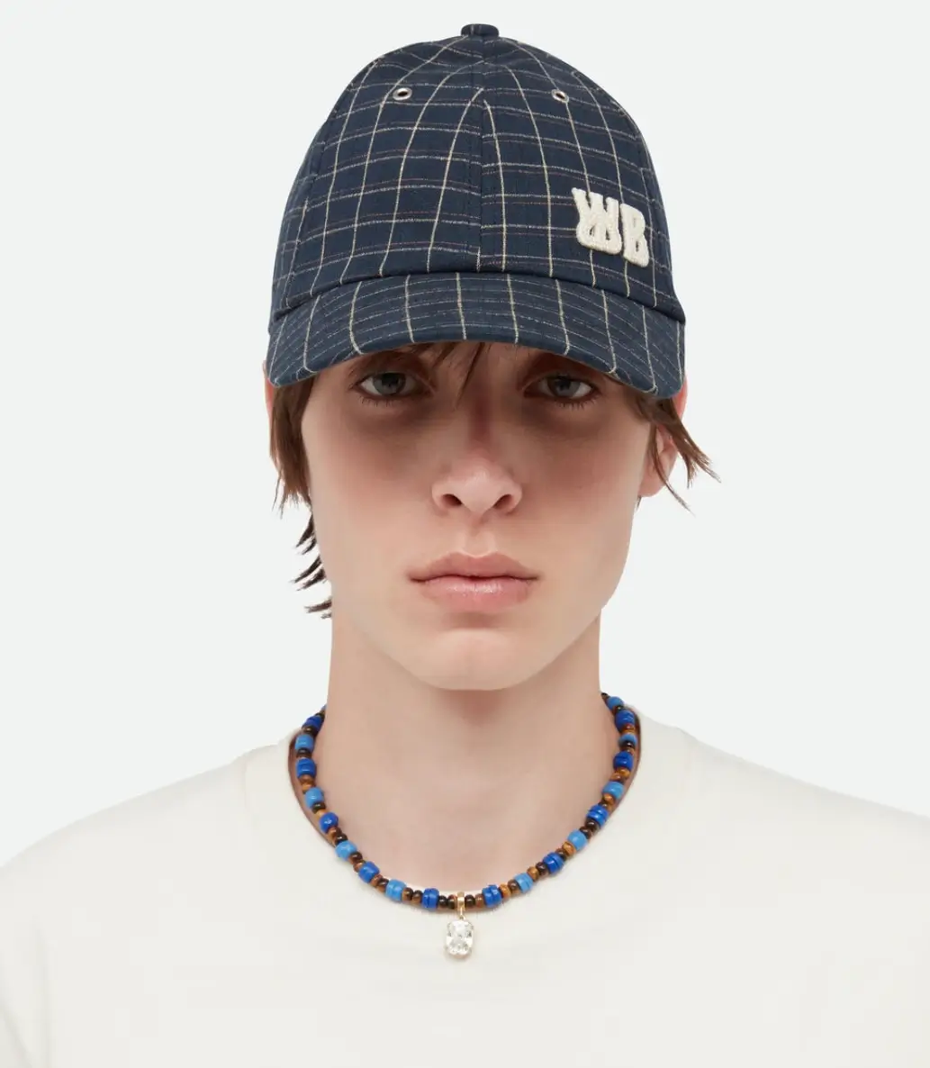 Portrait of a model wearing a navy check baseball cap with embroidered WB logo from the Wales Bonner Spring Summer 2026 collection, paired with a blue beaded necklace and white crewneck top against a neutral background