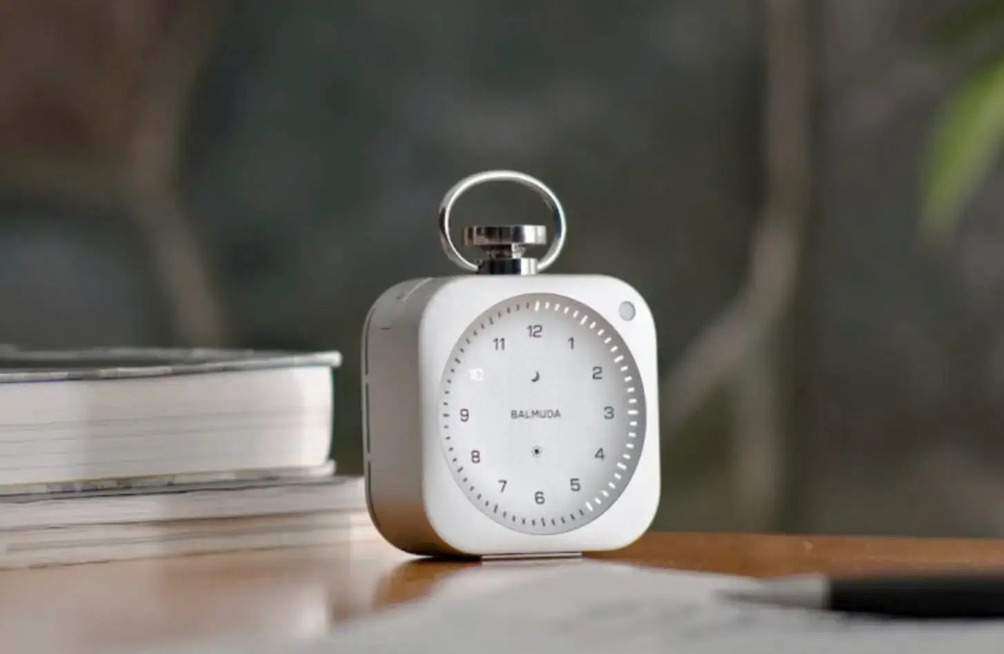 BALMUDA The Clock placed on a wooden desk beside stacked books in a softly lit interior, showcasing its minimalist aluminum body, illuminated dial, and refined, design-forward presence in a calm workspace setting