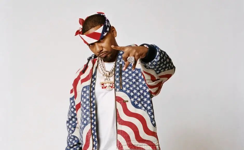 Portrait of Juelz Santana wearing a star-spangled American flag jacket and matching bandana, layered with gold chains, pointing toward the camera against a clean studio background