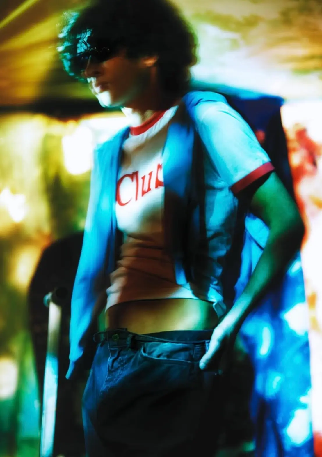 Blurred, cinematic portrait of a model in motion wearing a white ringer T-shirt with red trim and lettering, layered with a draped blue garment over the shoulders. The figure stands in a relaxed pose with one hand in the pocket of dark trousers, while oversized sunglasses and tousled hair add a sense of anonymity. Saturated neon hues—greens, yellows, and blues—bleed across the frame, creating a dreamlike, almost heat-hazed atmosphere that evokes movement, nightlife, and a softened, retro-futurist mood