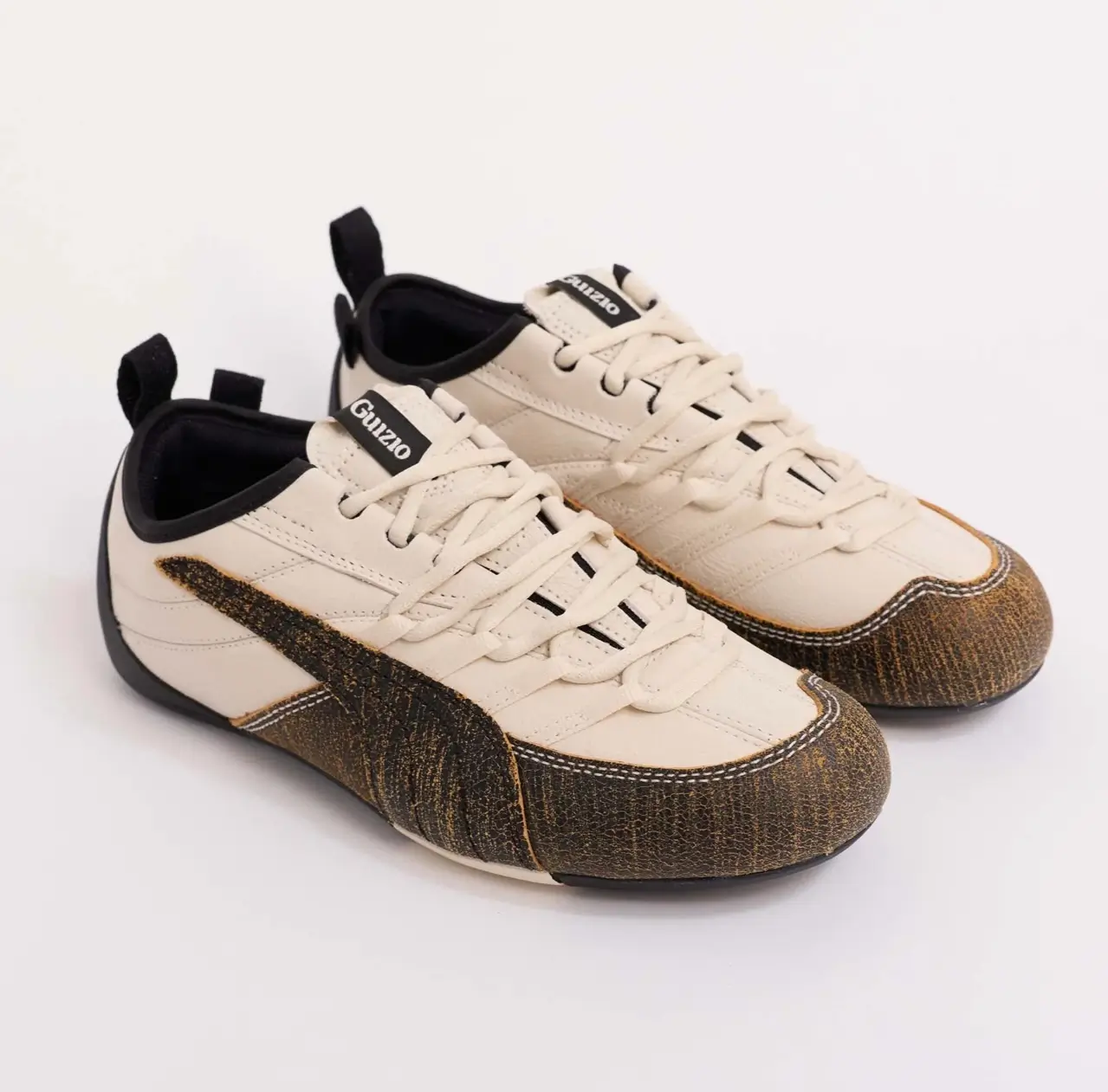 Three-quarter view of the PUMA x GUIZIO Klim Low shoe, showing cream leather uppers, tonal laces, black collar lining, Guizio-branded tongue tabs, and textured brown overlays extending into the Puma formstrip along the sides