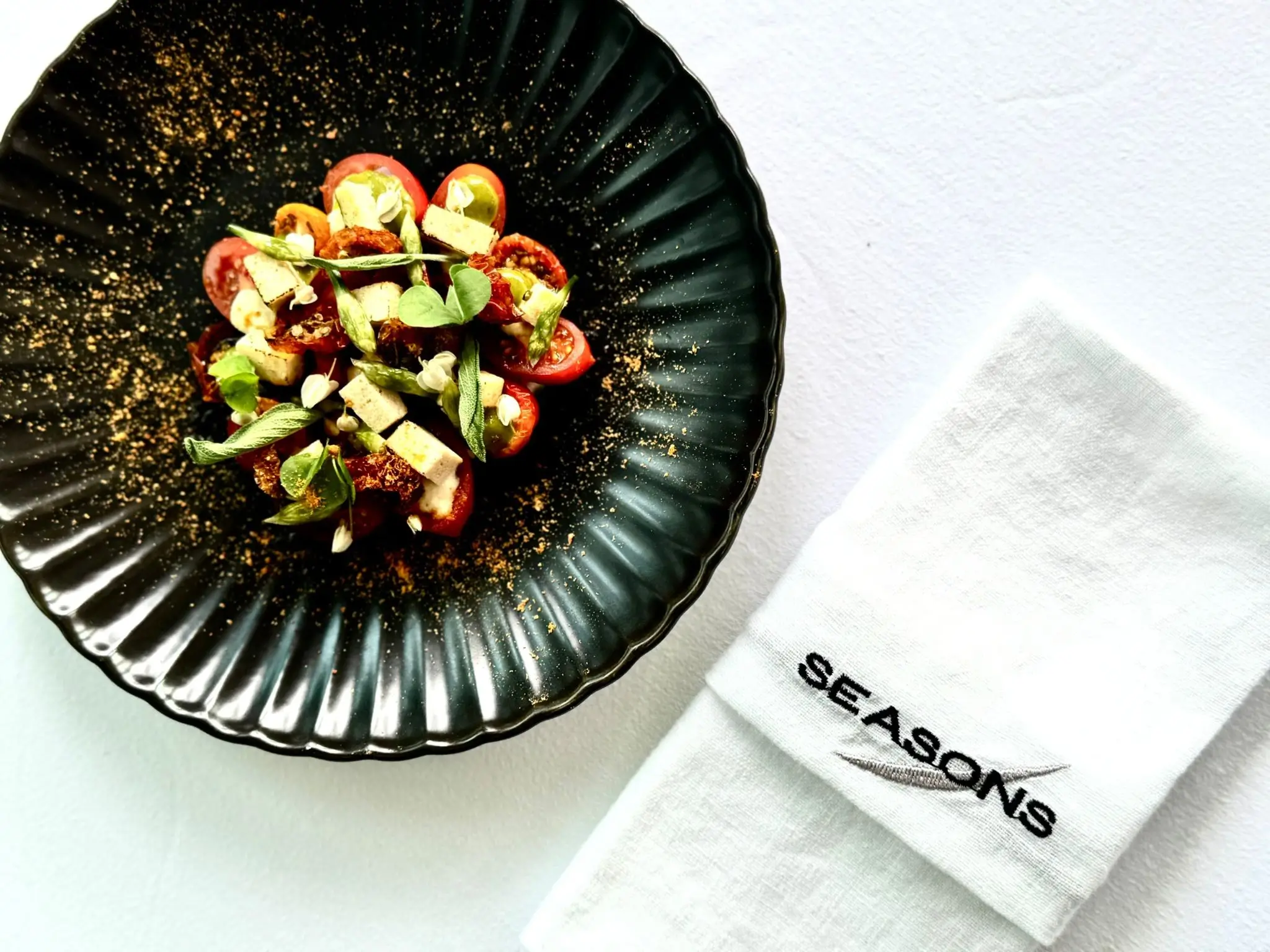 Top-down view of a refined plated dish at The SEASONS, featuring vibrant vegetables and delicate garnishes in a black textured bowl, accompanied by a folded white napkin embroidered with the SEASONS logo