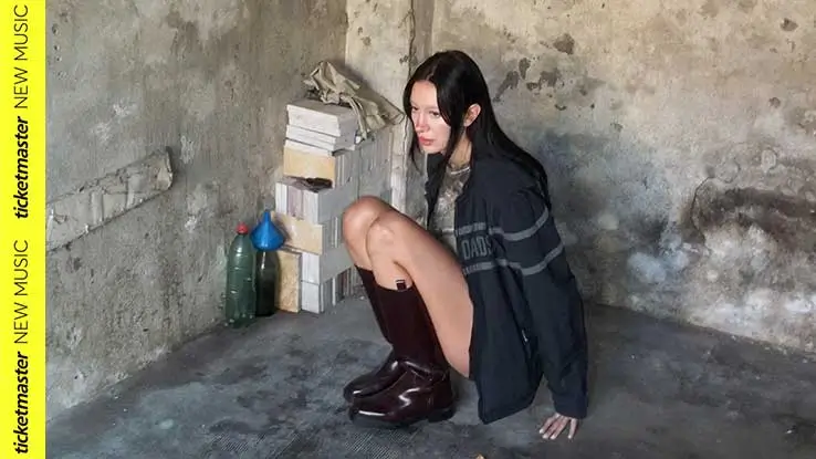 Julia Wolf crouching in a raw, concrete-walled room beside stacked books and bottles, wearing an oversized dark jacket and knee-high burgundy boots in a moody promotional photo for a new music feature