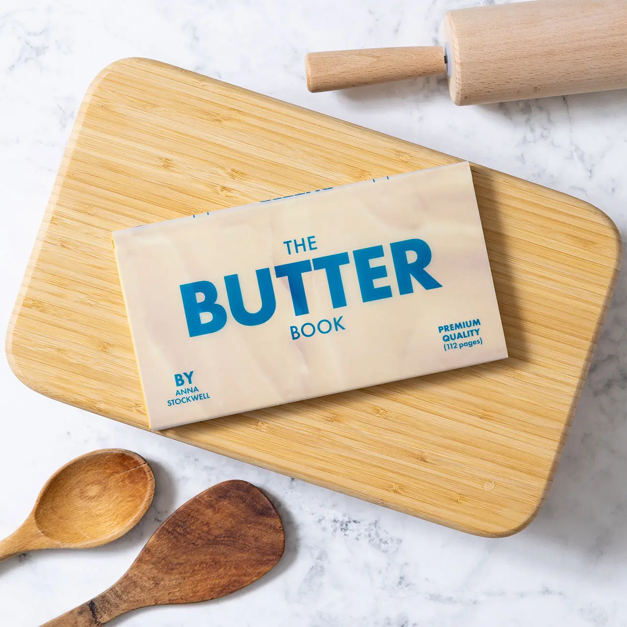 Cover of The Butter Book by Anna Stockwell placed on a wooden cutting board, featuring bold blue typography on a cream background, styled alongside a rolling pin and wooden spoons on a marble surface