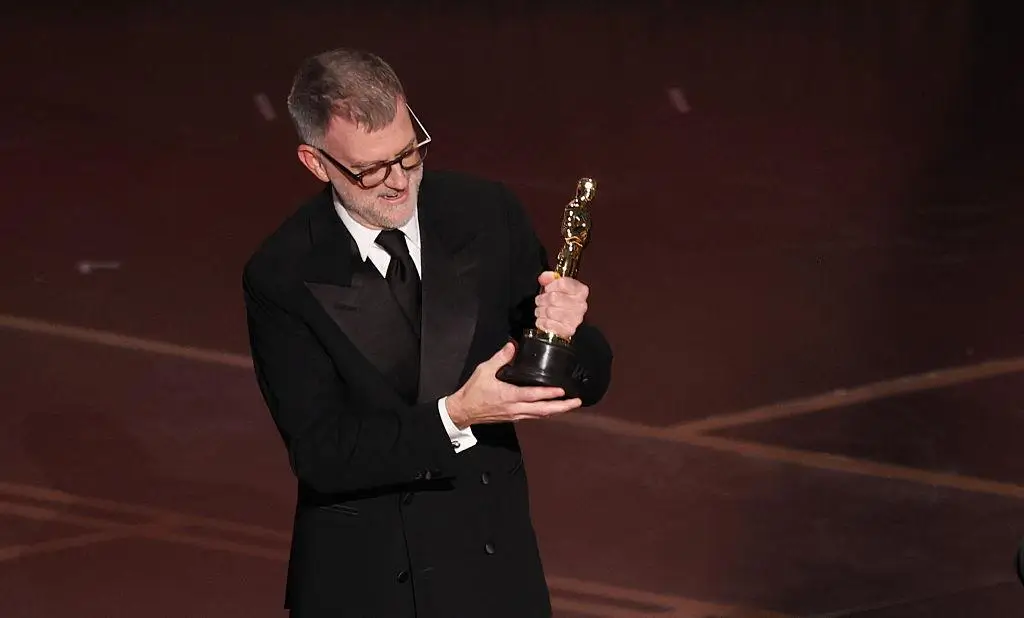 Paul Thomas Anderson holds the Academy Award on stage after winning Best Director at the Oscars, wearing a black tuxedo under warm stage lighting
