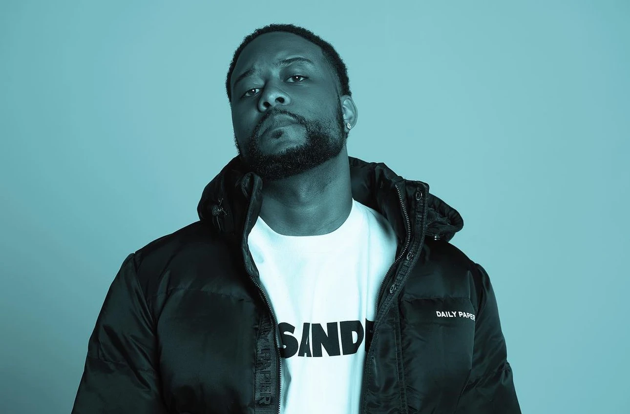 Portrait of music producer Supah Mario wearing a black Daily Paper jacket and white graphic T-shirt against a teal background, highlighting the South Carolina beatmaker known for trap and jazz experimentation