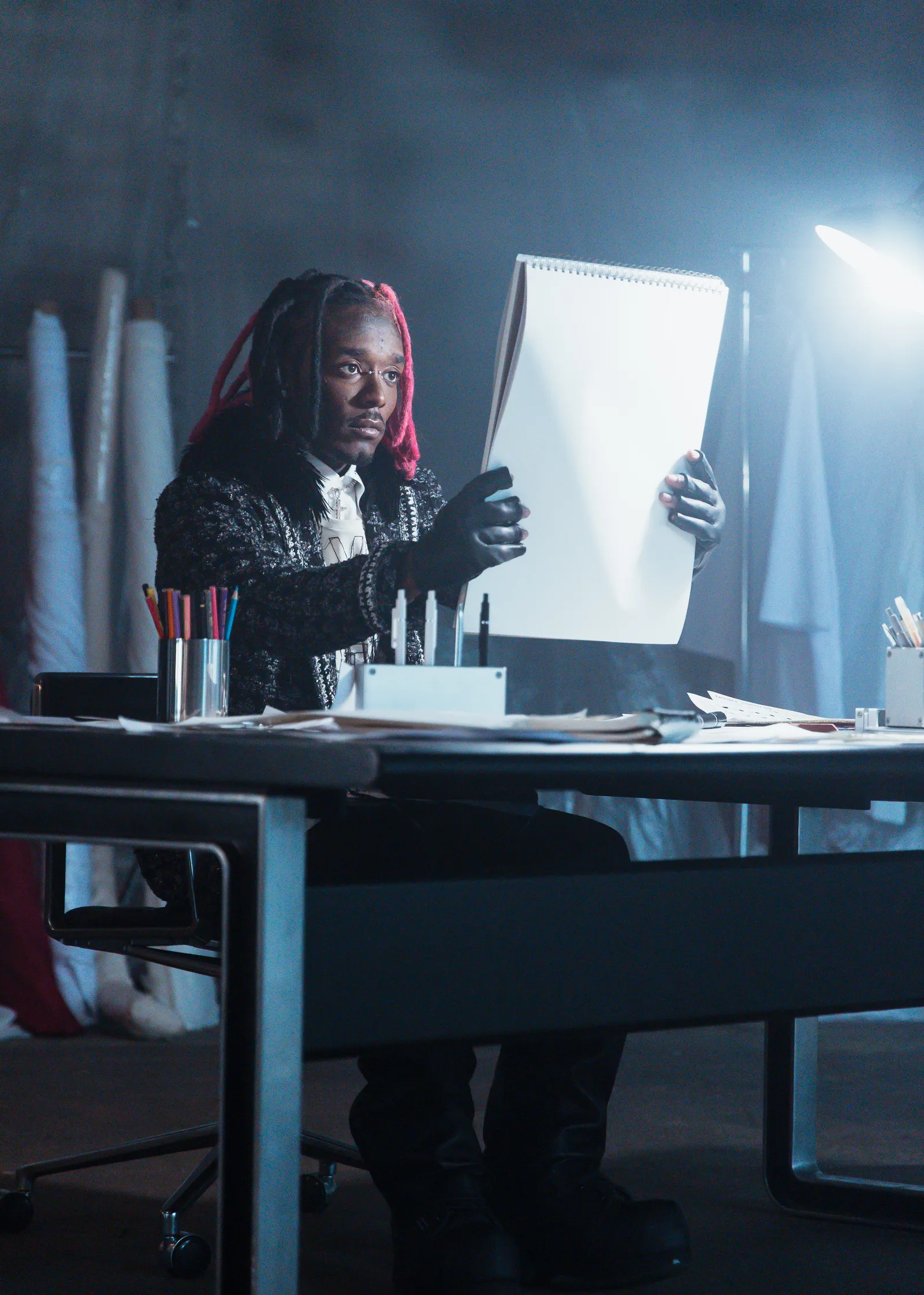 Lil Uzi Vert seated at a studio worktable, holding a large sketchpad under bright directional light, surrounded by drawing tools and papers in a moody, minimalist creative setting