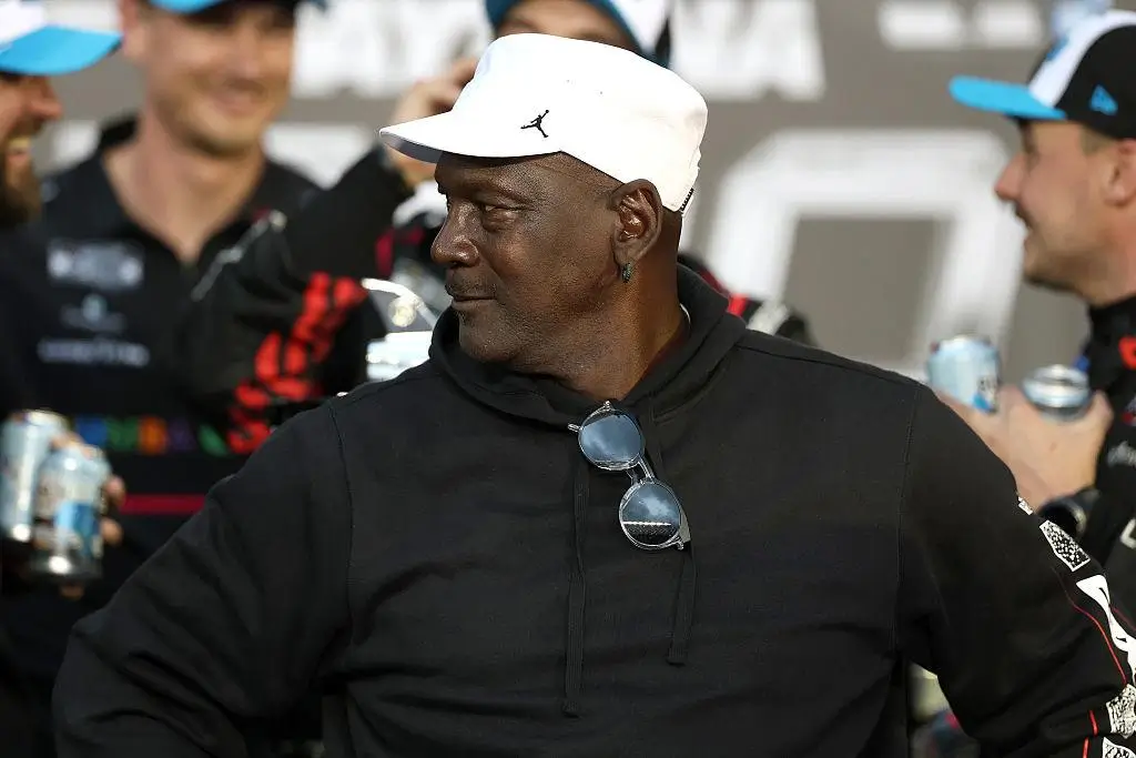 Michael Jordan wearing a white Jordan-branded cap and black hoodie, standing in a NASCAR team garage area, looking to his left while crew members in racing uniforms hold drinks in the background