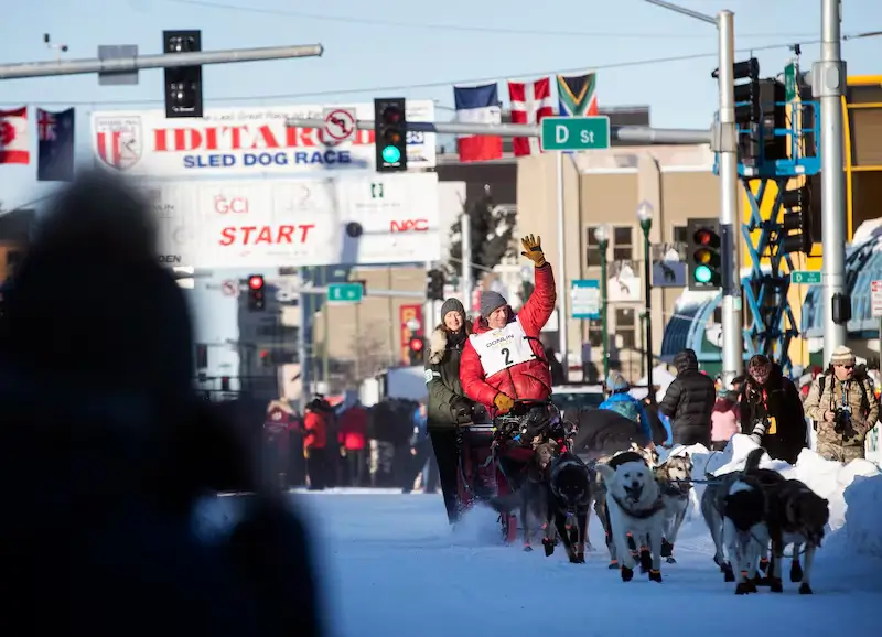 Musher wearing bib number 2 waves to spectators while guiding a team of sled dogs through downtown Anchorage during the ceremonial start of the 2026 Iditarod Trail Sled Dog Race, with the start banner, traffic lights, and city buildings visible in the background