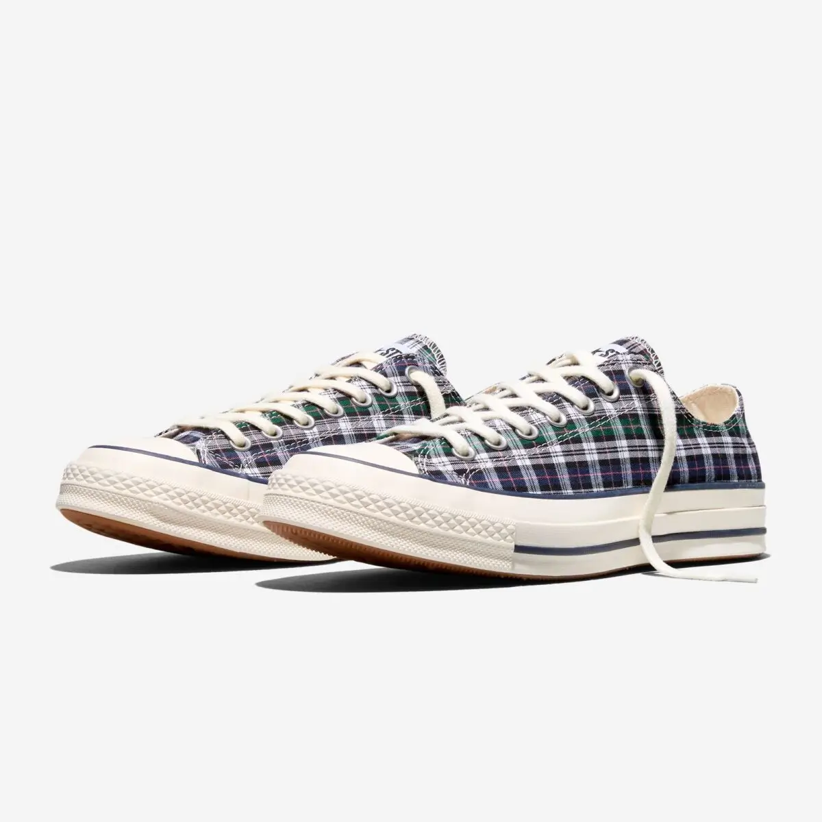 Angled front view of Converse Chuck 70 low-top shoe in a navy, green, and white plaid canvas, featuring cream laces, textured rubber toe caps, and off-white vintage-style midsoles with a thin navy stripe; the paired presentation emphasizes the classic silhouette and understated collaboration with Noah