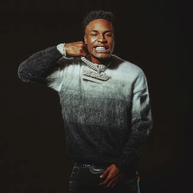 Studio portrait Mudbaby Ru of a rapper wearing a grey gradient sweater and diamond chain, holding the pendant up while showing a grill, set against a dark background
