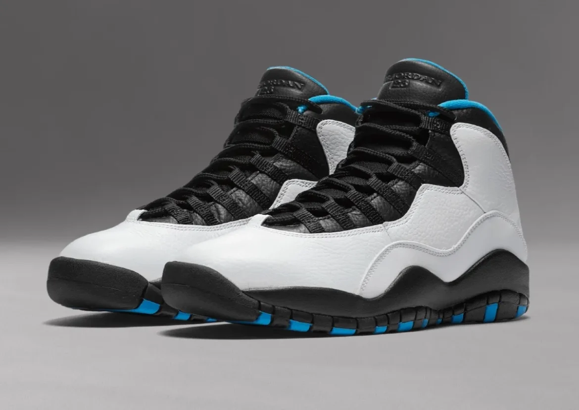 Angled front three-quarter view of a pair of Air Jordan 10 “Powder Blue” sneakers on a neutral grey background, showing white tumbled leather uppers, black leather tongues and eyestays with black rope laces, sculpted black midsoles, and bright powder blue accents peeking through the outsole and collar lining