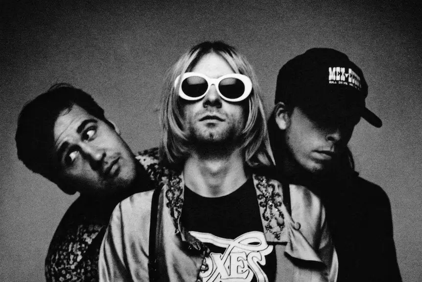 Kurt Cobain wearing white oval sunglasses standing between bandmates Dave Grohl and Krist Novoselic in a black-and-white Nirvana portrait