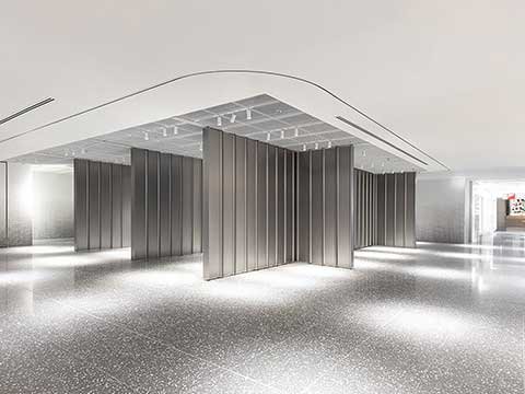 Minimalist underground retail installation featuring a grid of tall, brushed metal panels arranged in a sculptural formation beneath a recessed ceiling with linear lighting, set against a polished speckled floor and expansive open space, evoking a quiet, gallery-like environment