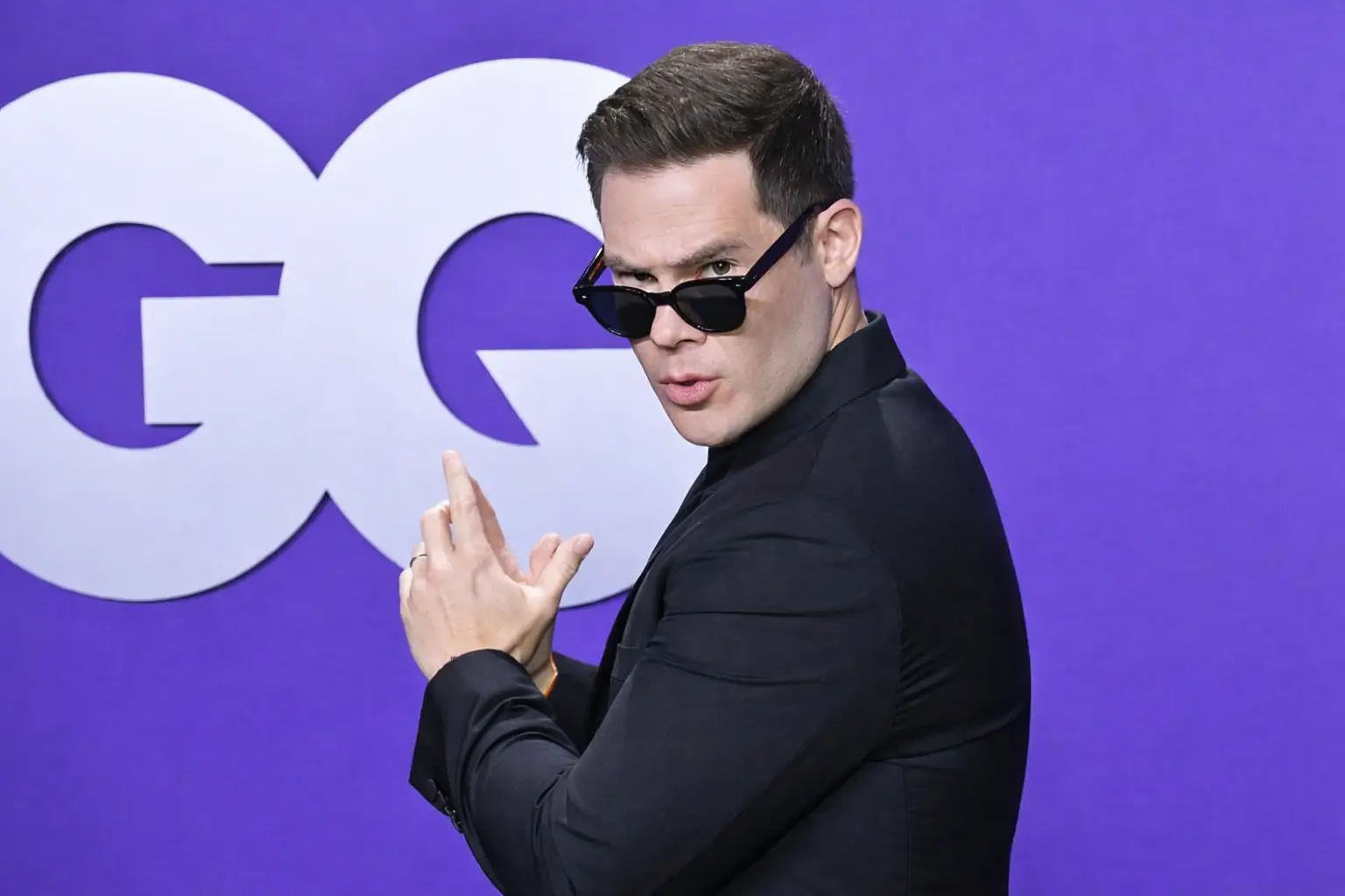Adam DeVine tailored black suit and dark sunglasses poses with a playful finger-gun gesture against a smooth purple backdrop featuring GQ large white lettering, captured in evenly diffused lighting that minimizes shadows and emphasizes a clean, polished appearance