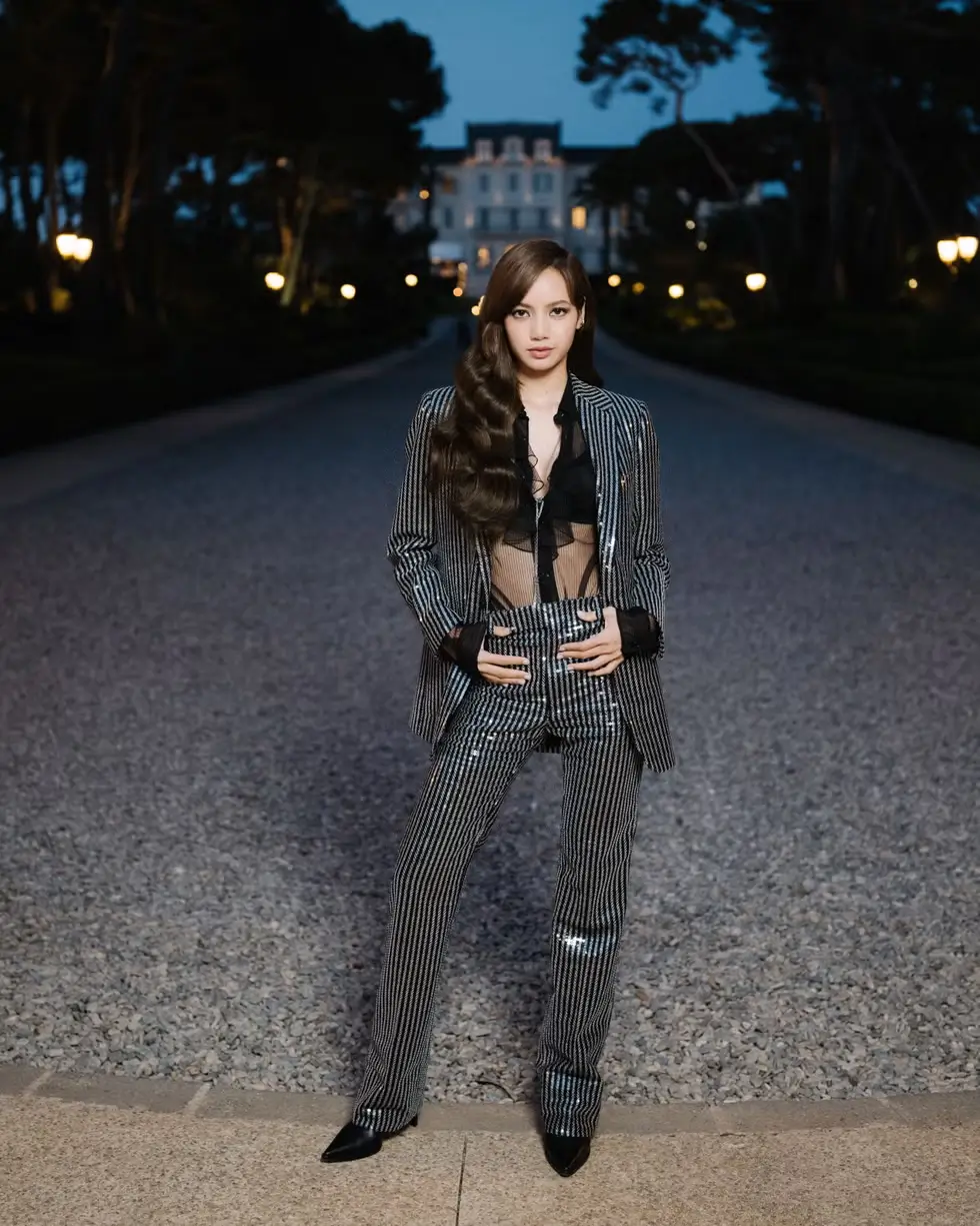 Lisa stands centered on a gravel pathway at dusk, wearing a sharply tailored pinstripe suit layered over a sheer black top, with a softly lit villa and trees framing the background in a cinematic evening setting