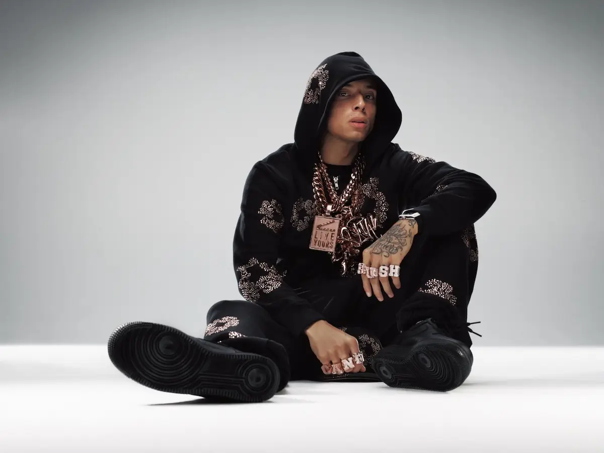 Studio portrait of a Central Cee seated on the floor against a neutral grey backdrop, wearing a black embellished hoodie and matching pants, layered heavy chain necklaces, rings, and black sneakers, with a relaxed, confident pose and hood partially covering his head