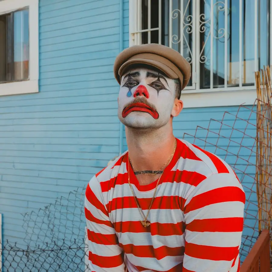 Portrait of Jacob Dietrich wearing white clown makeup with black eye detailing and a red nose, dressed in a red-and-white striped shirt and brown cap, seated outdoors against a pale blue building