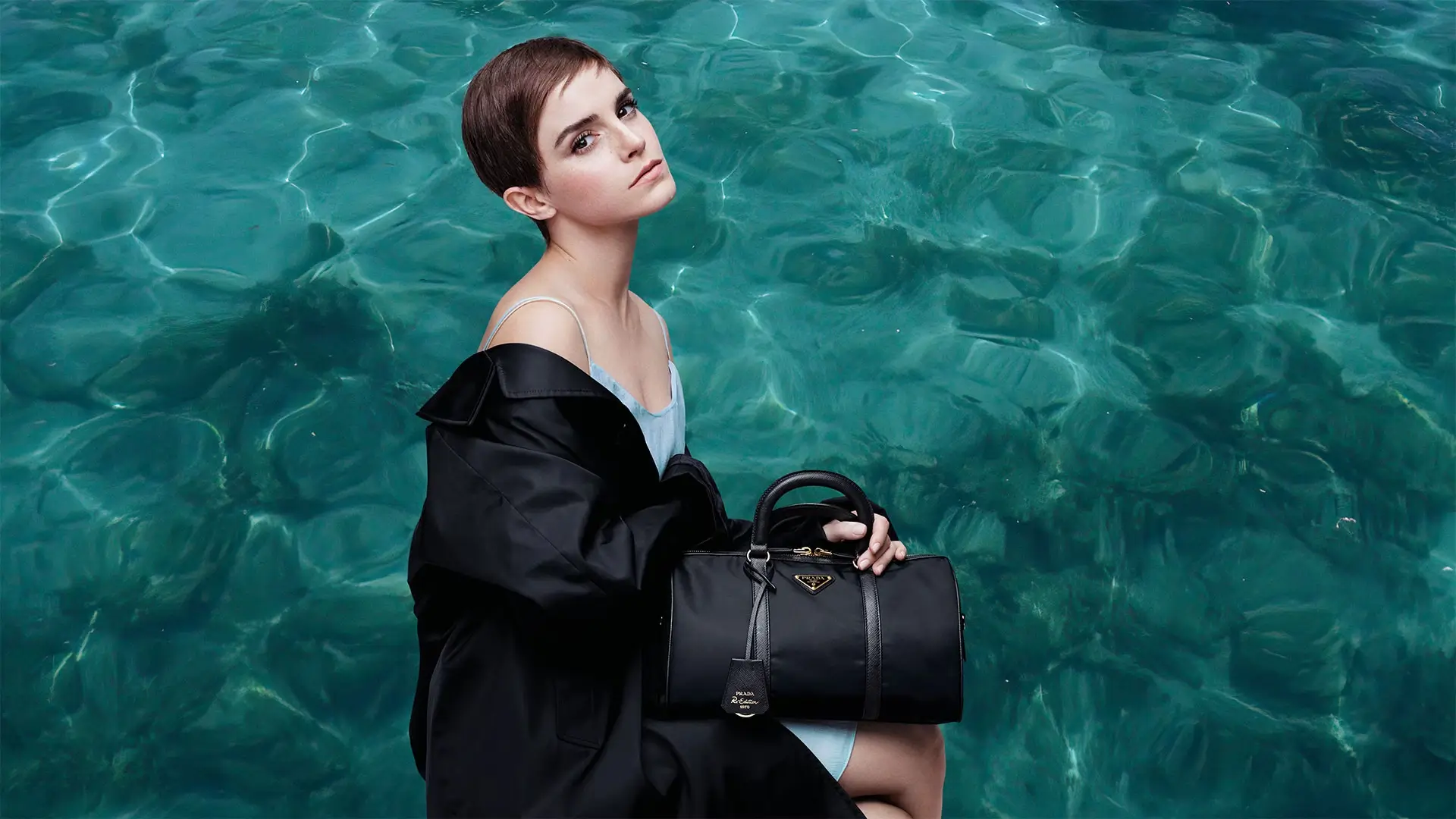 Model seated against a backdrop of clear turquoise water, wearing a black Prada jacket and holding a black Prada Re-Nylon handbag with top handles and logo plaque
