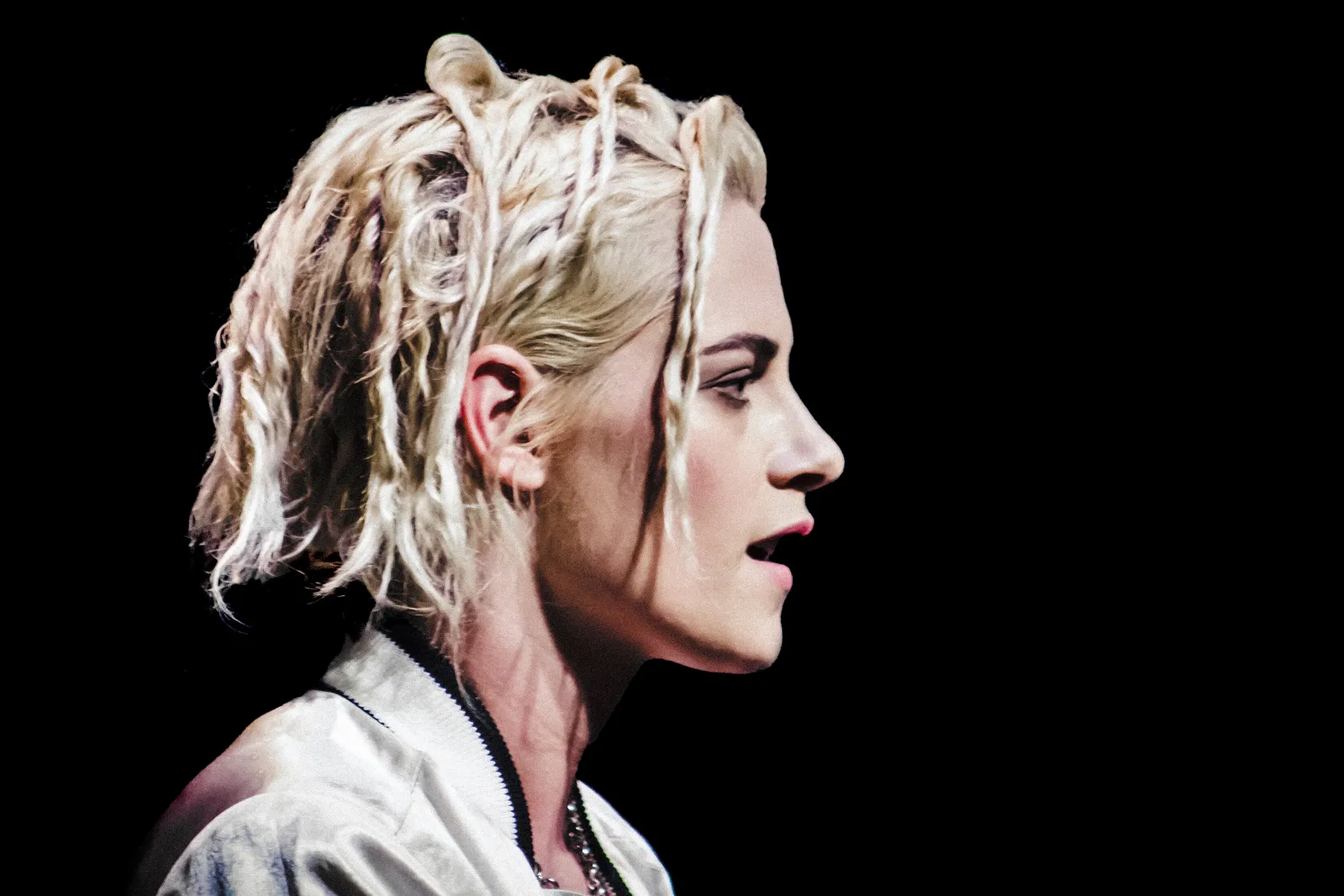 Side-profile portrait of Kristen Stewart with short bleached hair styled in textured strands, wearing a light jacket against a black background, her face lit dramatically to emphasize sharp features and a focused expression