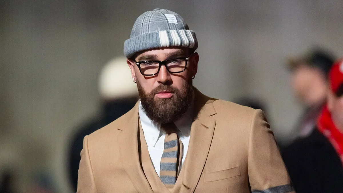 Close-up of Travis Kelce wearing a camel-colored tailored suit with a striped tie, clear-framed glasses, and a grey knit beanie, walking outdoors with a softly blurred crowd in the background