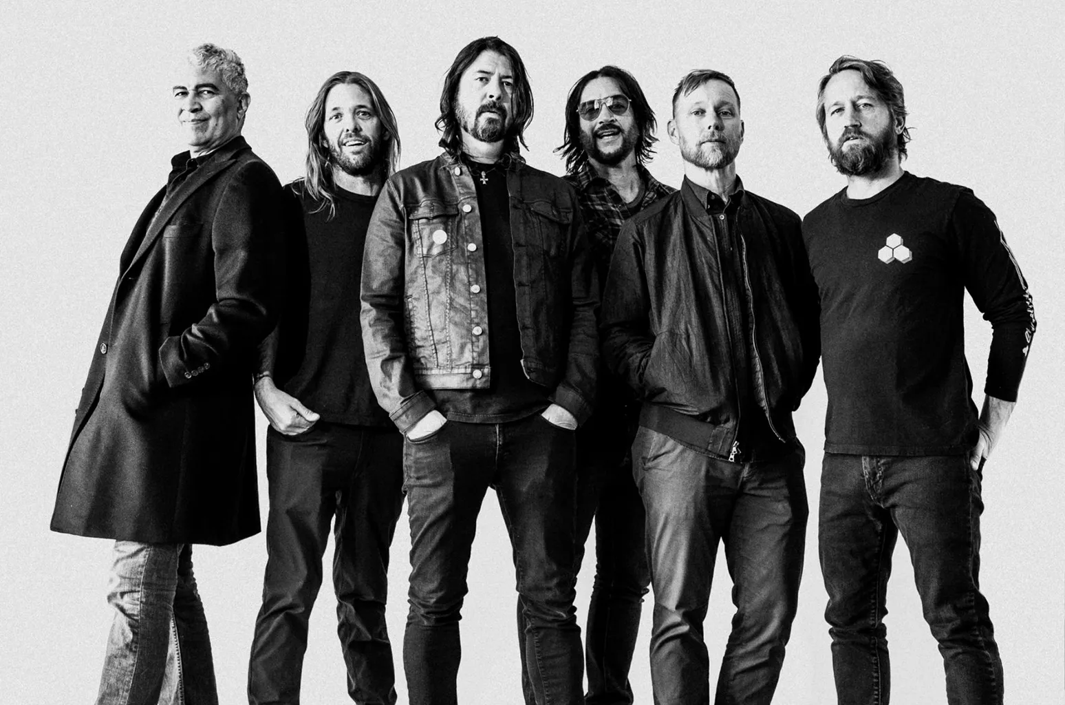 A black-and-white press photo shows the members of Foo Fighters standing together in a studio setting, dressed in casual dark clothing, with Dave Grohl centered among the group, conveying a unified, understated rock band presence