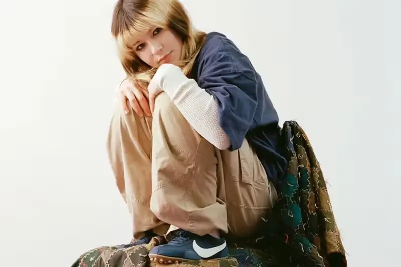Alysa Liu seated in a relaxed pose wearing layered neutral-toned Nike apparel, including loose khaki pants and a navy top, paired with dark Nike sneakers featuring a white Swoosh, set against a clean studio background