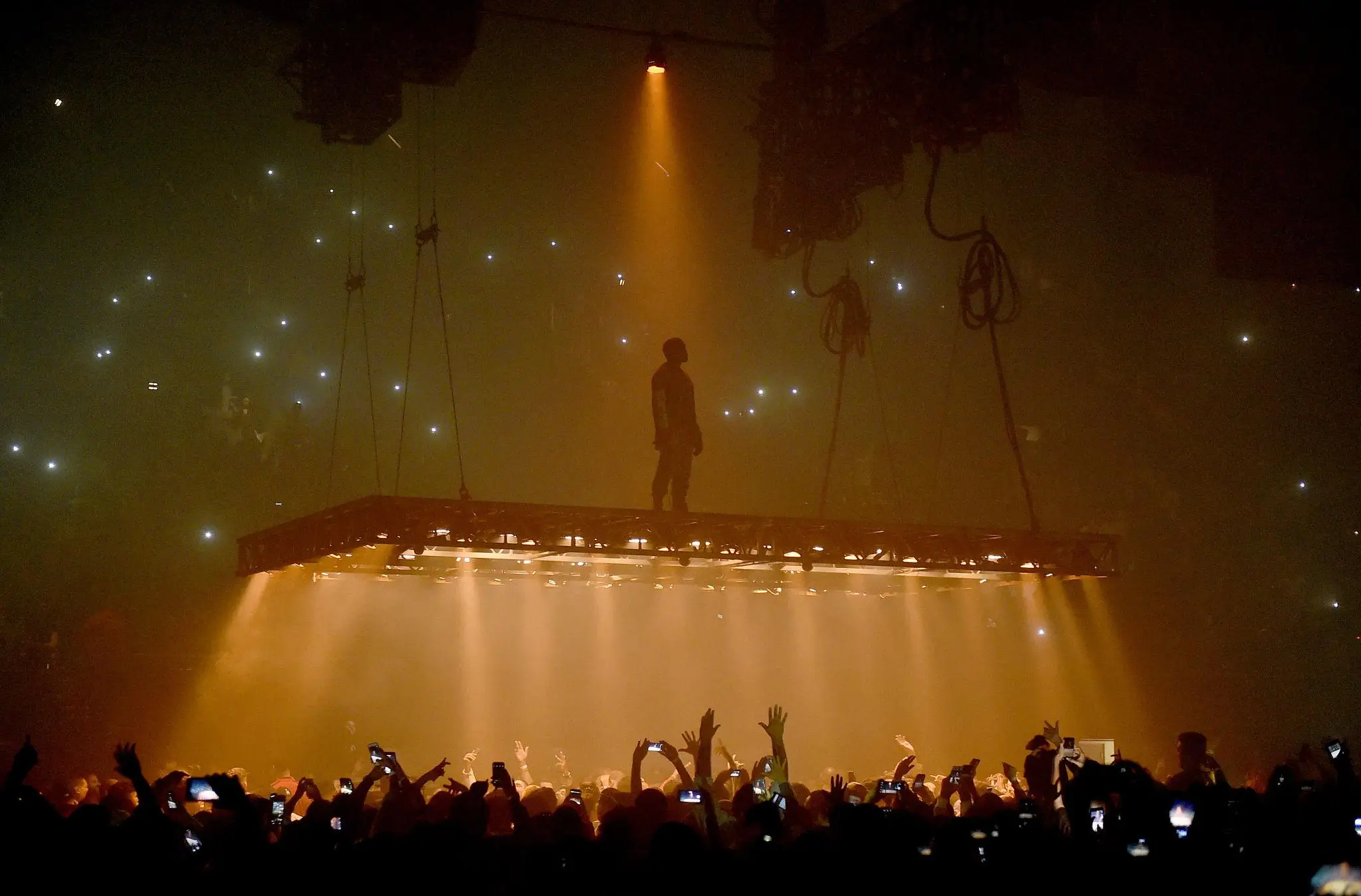 Kanye West stands silhouetted on a suspended floating stage platform above a massive crowd during a concert performance, illuminated by dramatic amber lighting while thousands of fans below hold up smartphones in a dark arena