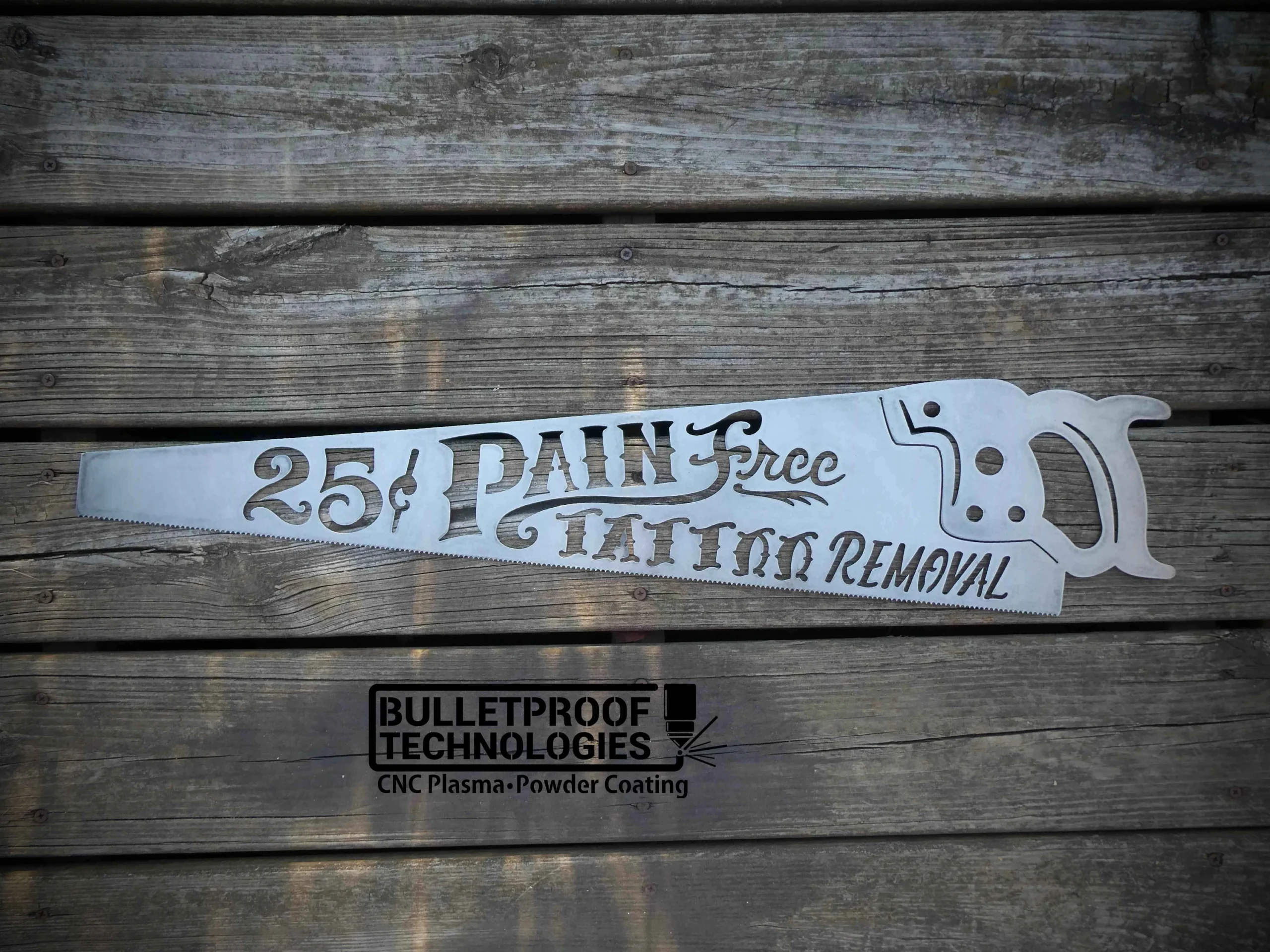 Metal saw-shaped sign reading “25¢ Pain Free Tattoo Removal” placed on weathered wooden boards, featuring decorative typography and branding from Bulletproof Technologies CNC Plasma Powder Coating