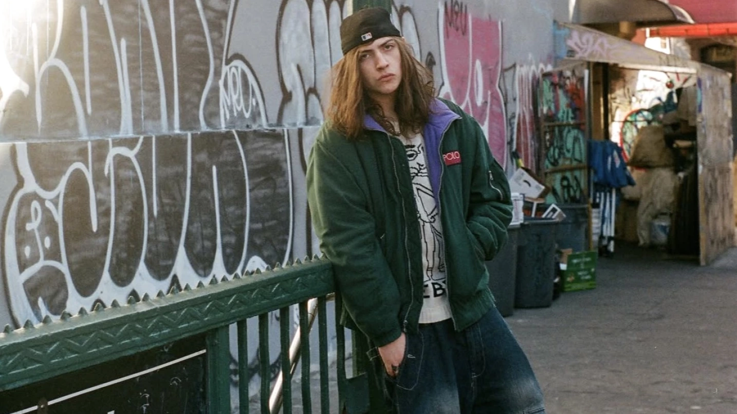 Sko with long hair and a black cap stands against a graffiti-covered wall, wearing a green jacket layered over a graphic T-shirt, hands tucked casually as he leans beside a metal railing in an urban alley filled with street art and scattered objects