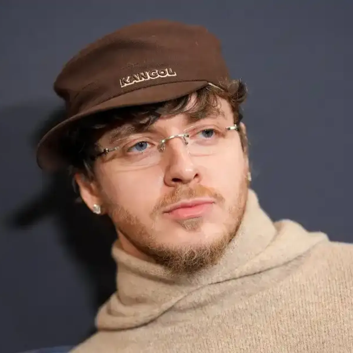 Portrait of rapper Jack Harlow wearing a brown Kangol cap, rimless glasses, and a beige knit hoodie, photographed at a film premiere event