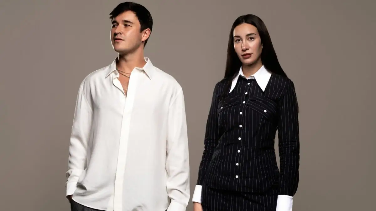John Summit wearing a white button-down shirt standing beside Julia Wolf in a black pinstripe dress with white collar and cuffs against a neutral studio background, promoting their collaboration “With Me.”