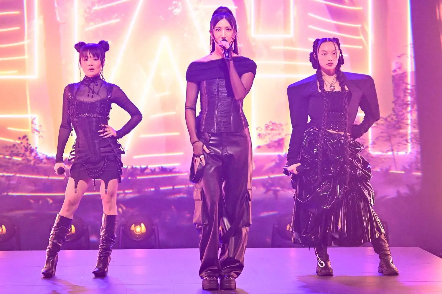 Three K-pop performers stand center stage under glowing pink and amber lights, dressed in coordinated black avant-garde outfits, with the lead singer holding a microphone while the others pose confidently beside her