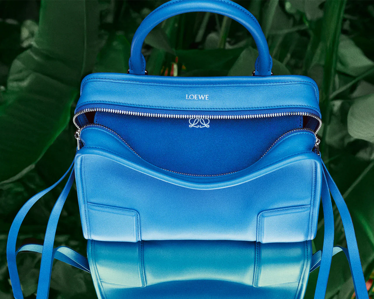 Close-up of a cobalt blue Loewe Amazona 180 top-handle bag with curved front panel and silver zipper detail against a lush green foliage background