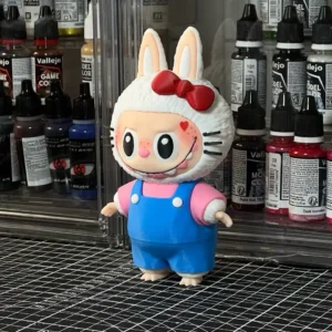Custom Labubu designer toy styled with Hello Kitty elements, featuring a white bunny hood with a red bow and whiskers, wearing blue overalls and standing on a craft table surrounded by model paint bottles