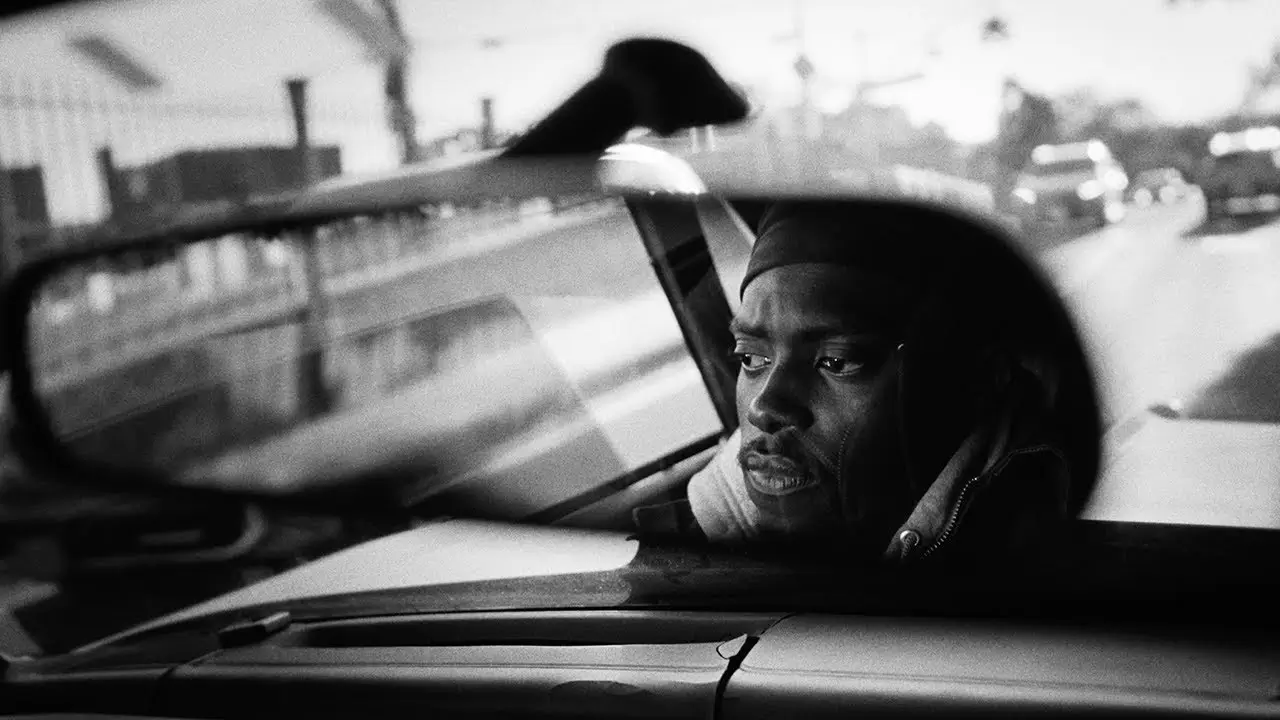 Black-and-white cinematic frame of Niontay in a car, seen through the rearview mirror, gazing out with a contemplative expression as blurred urban streets pass behind him