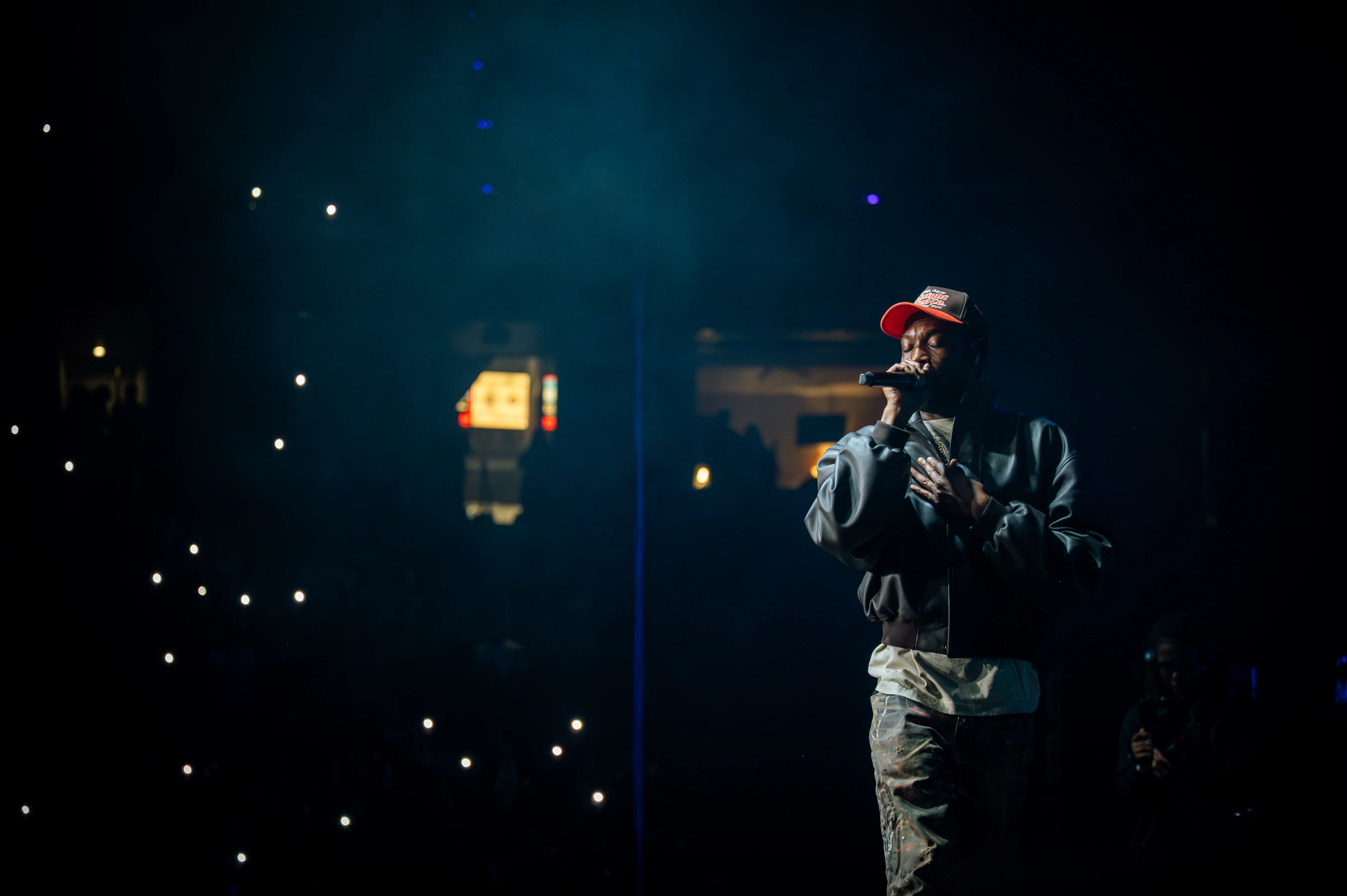 Meek Mill performs live on stage at Clover Fest, illuminated by a single overhead spotlight, wearing a red cap and layered streetwear while holding a microphone close, with a darkened crowd behind him lit by scattered phone lights