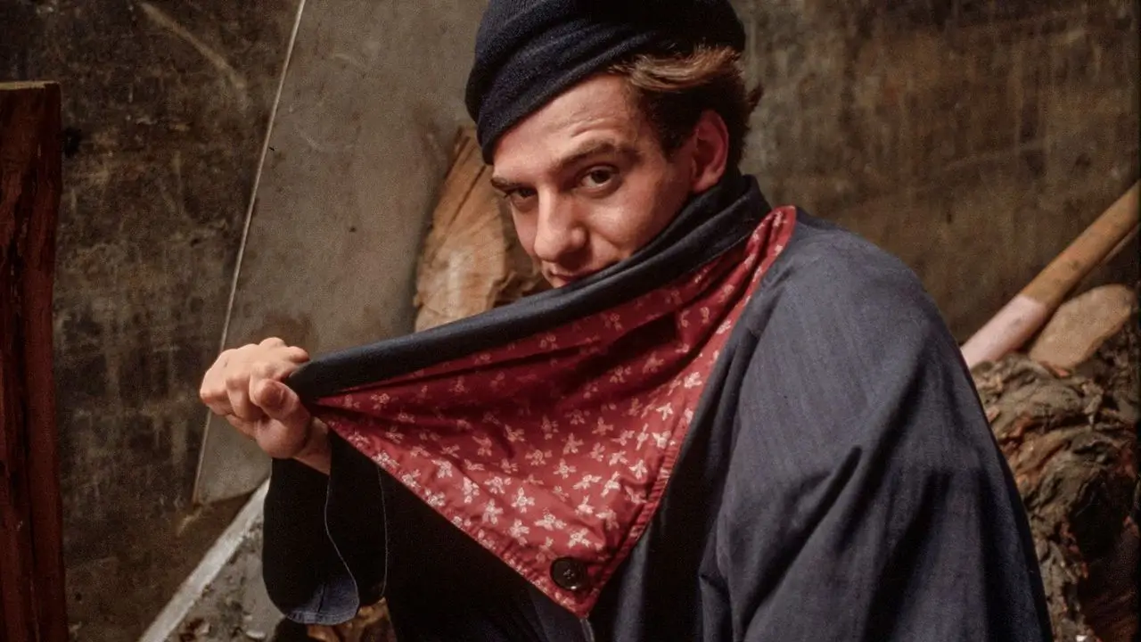 John Galliano wearing a dark beret and oversized coat pulls up a red patterned scarf to partially cover his face, posing in a rustic, dimly lit interior with textured walls and wooden elements in the background