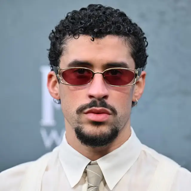 Bad Bunny wearing red-tinted sunglasses and a beige shirt with tie, posing for a portrait at a film premiere event