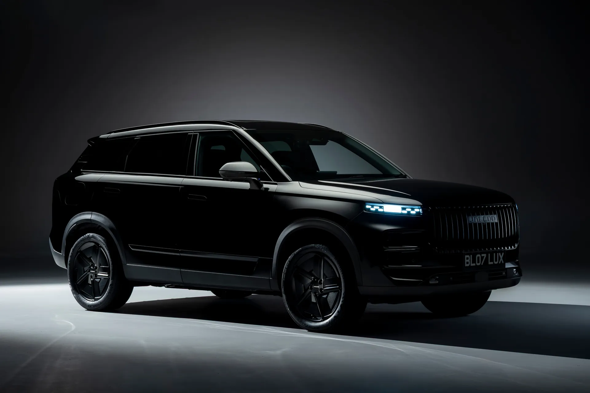 Three-quarter front view of the Jaecoo 7 SUV in a dark studio, finished in black with illuminated LED headlights, a vertical-slat grille, and shadowed body lines emphasizing its boxy, refined silhouette
