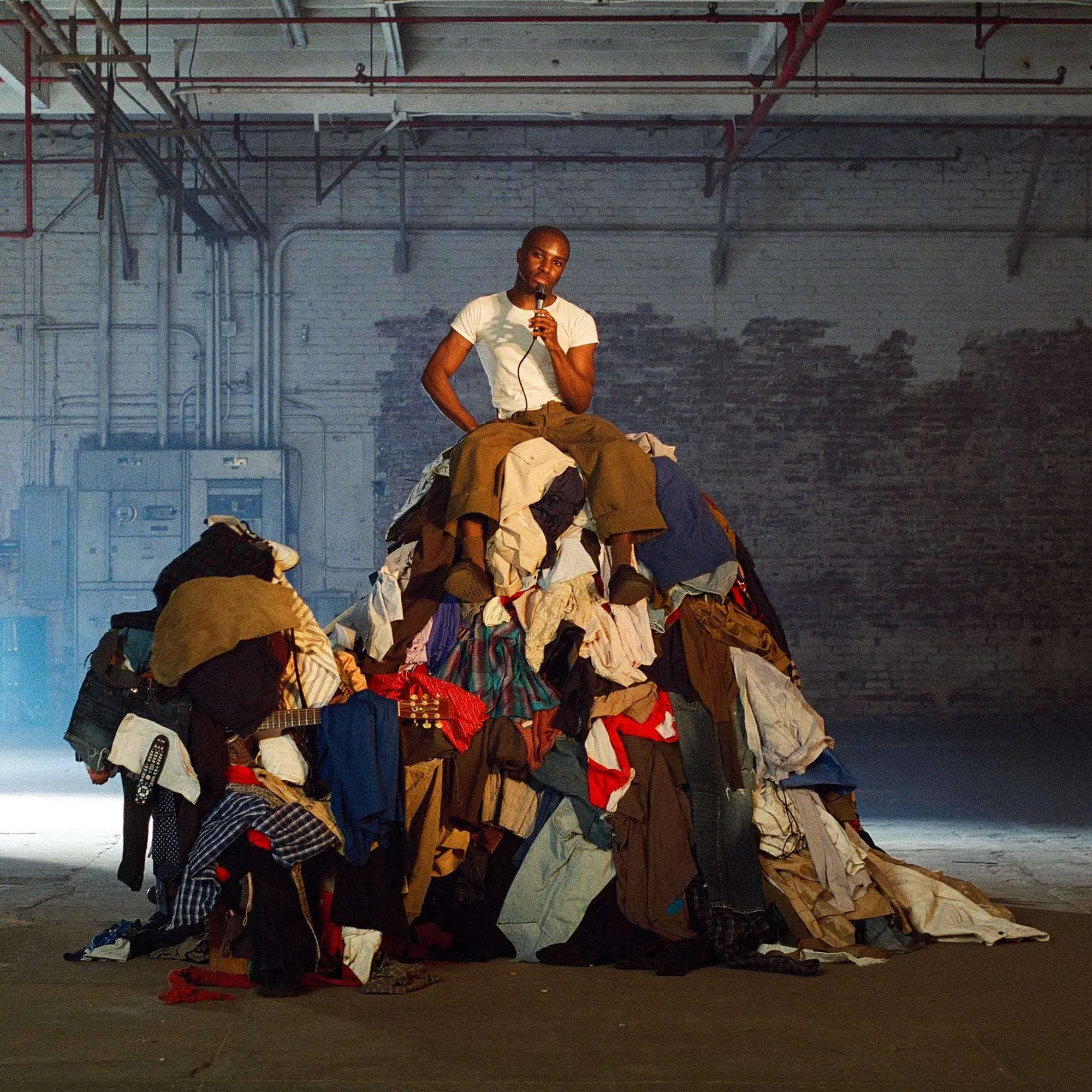 rum.gold seated atop a large mound of mixed clothing in an industrial warehouse, holding a microphone and wearing a fitted white T-shirt and brown trousers, with exposed pipes and brick walls creating a raw, intimate performance setting