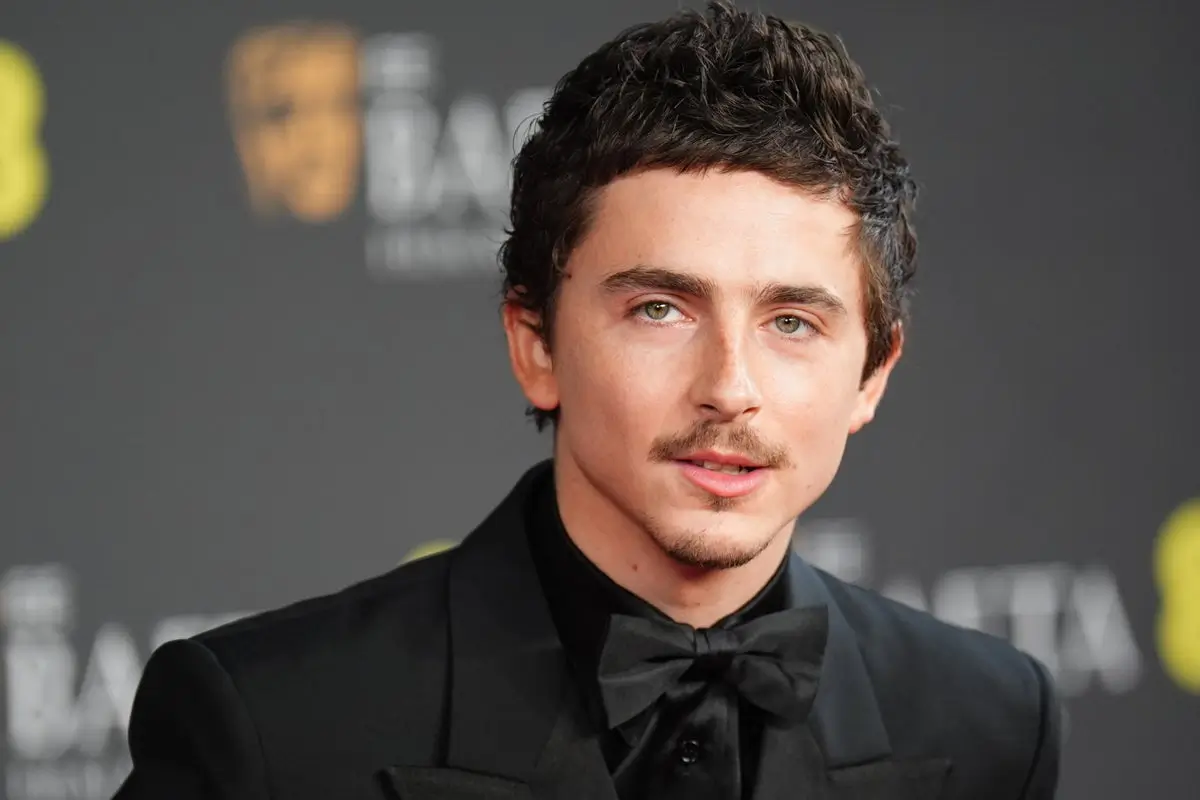 Timothée Chalamet wearing a black tuxedo and bow tie at a BAFTA Film Awards red carpet event, posing in front of a dark backdrop with event branding