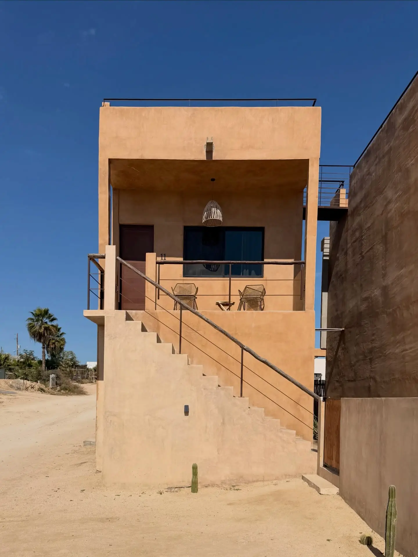 Minimalist desert villa in Cabo San Lucas with warm sand-toned stucco facade, exterior staircase, and shaded balcony seating under a clear blue spring sky, surrounded by arid landscape and sparse cacti
