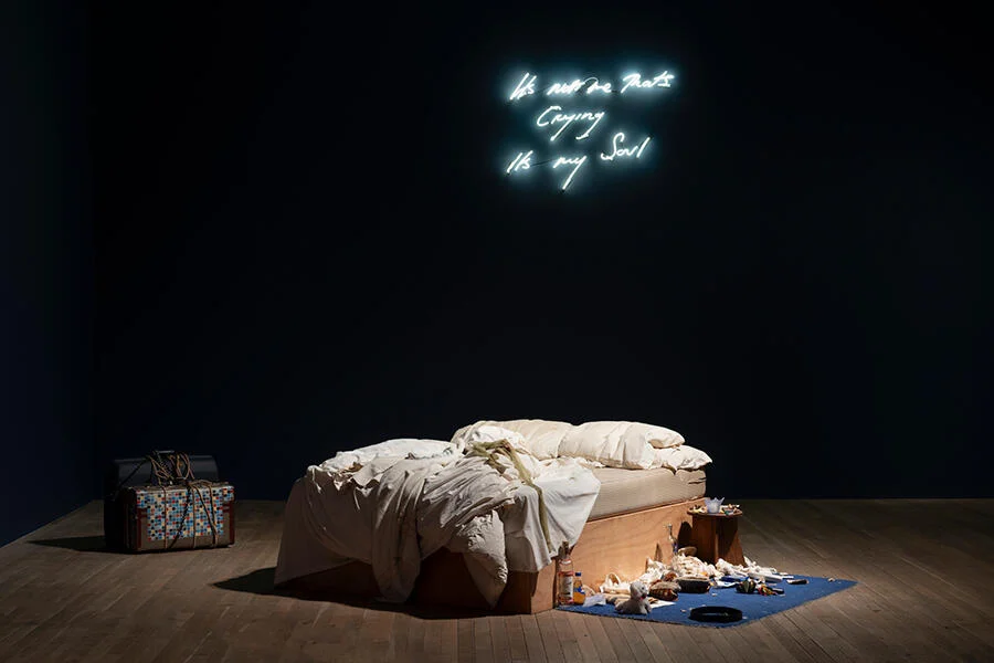 Installation view of Tracey Emin’s My Bed (1998) featuring a rumpled, unmade bed surrounded by personal items—bottles, clothing, and scattered belongings—set beneath a glowing handwritten neon text reading “It’s Not Me That’s Crying It’s My Soul” (2001) against a dark gallery wall