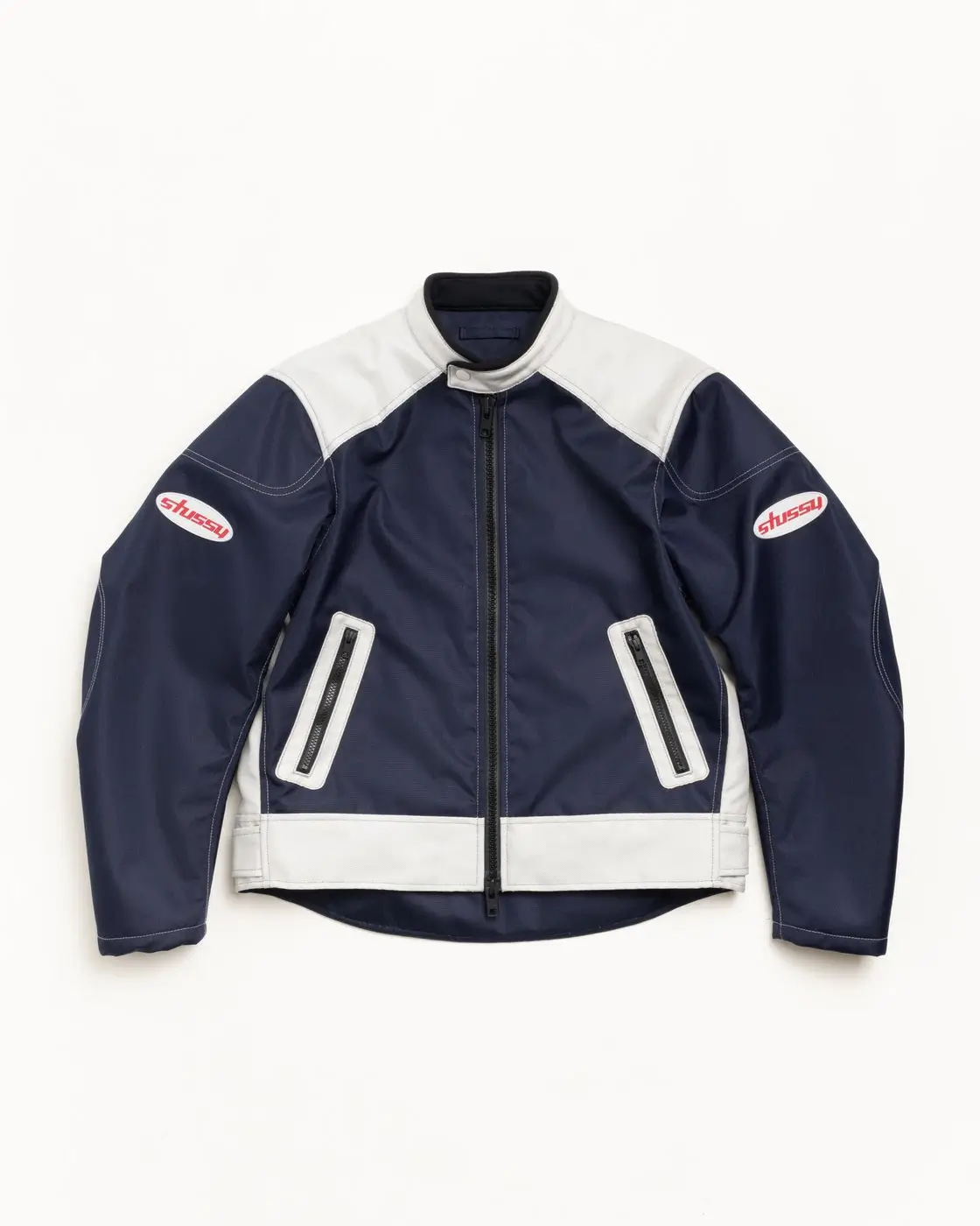 Navy and cream racing-style zip jacket with contrast shoulder panels, zippered pockets, and red Stüssy logo patches on both sleeves