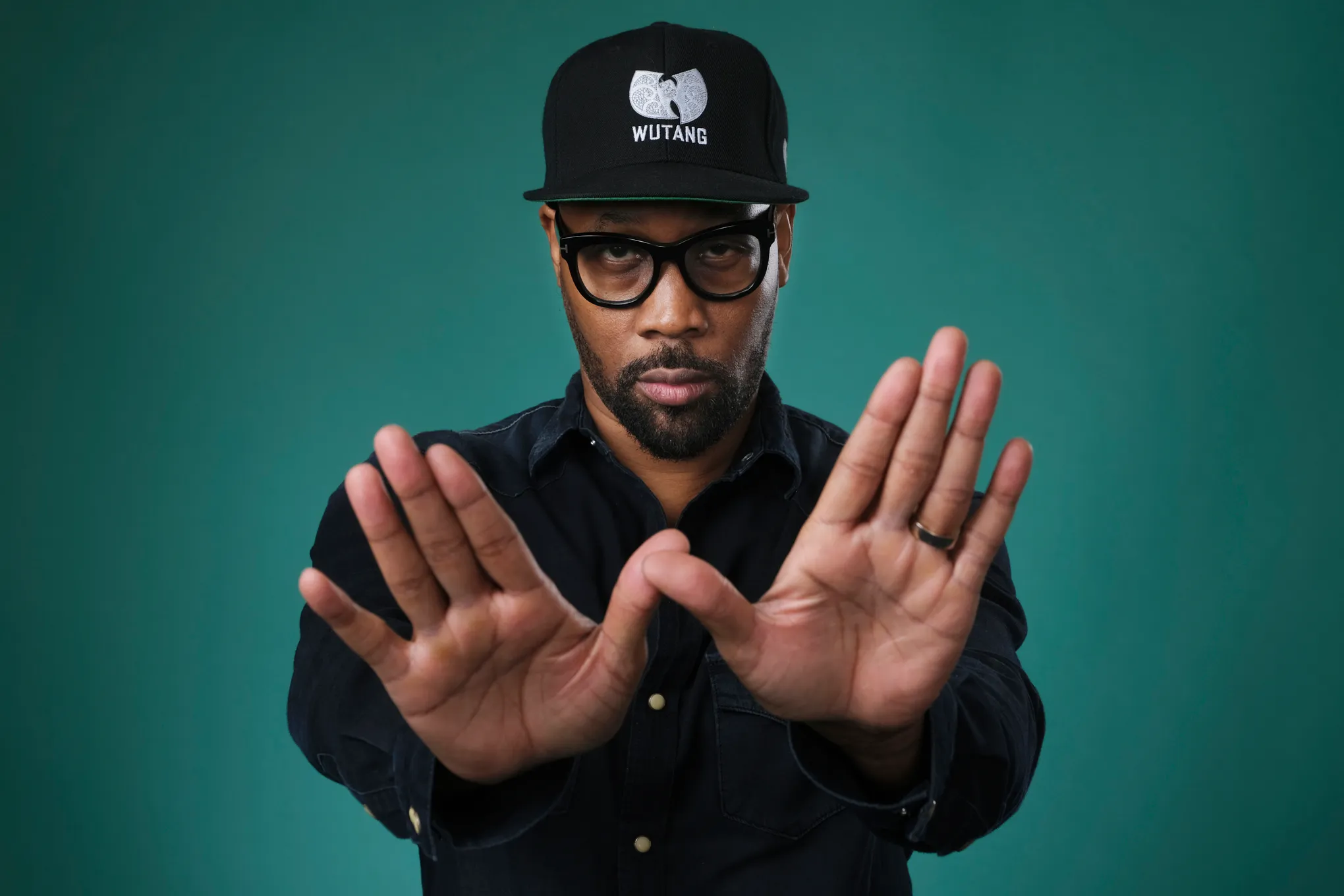 RZA poses against a teal background wearing a black Wu-Tang cap and glasses, holding both hands forward to form the iconic “W” hand sign associated with Wu-Tang Clan