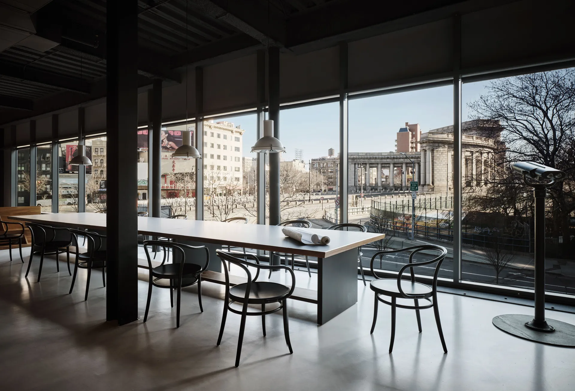 A bright studio workspace with long communal tables and black chairs set beside floor-to-ceiling windows, overlooking a city street and a neoclassical building, with natural light filling the interior