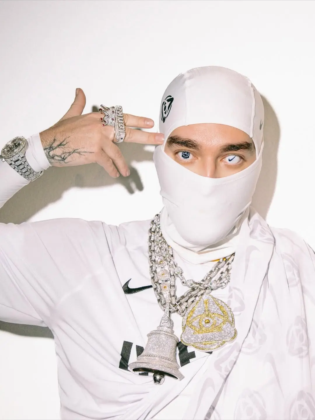 Yeat masked figure in white Nike balaclava and matching outfit wears diamond chains and rings, posing with hand gesture against a clean studio background
