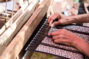 Artisan carefully weaving patterned textile on a traditional wooden handloom, guiding threads by hand as geometric fabric design emerges