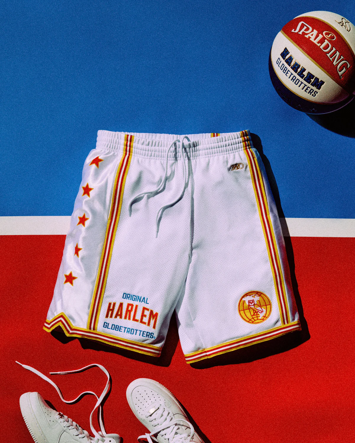 OVO x Harlem Globetrotters white basketball shorts with red and gold trim, star detailing, and “Original Harlem Globetrotters” branding, styled with a Spalding basketball and white shoe against a red, white, and blue backdrop